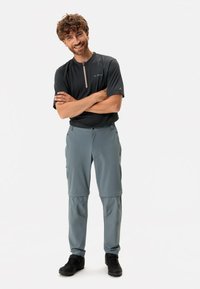 Heren sportoutfit bestaande uit een zwart rits kortmouwtop en grijze convertibele broek met zakken, gecombineerd met zwarte schoenen.