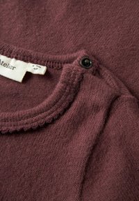 Burgundowy sweter z okrągłym dekoltem, z falbaniastym wykończeniem i czarnym guzikiem w pobliżu metki. Miękka tekstura. Zawiera metkę rozmiaru.