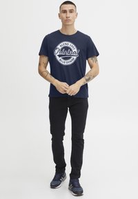 Mørkeblå bomull t-skjorte med hvit sirkulær "Original Blend 1993 Blue Denim" trykk, parret med svarte bukser og grå sneakers.