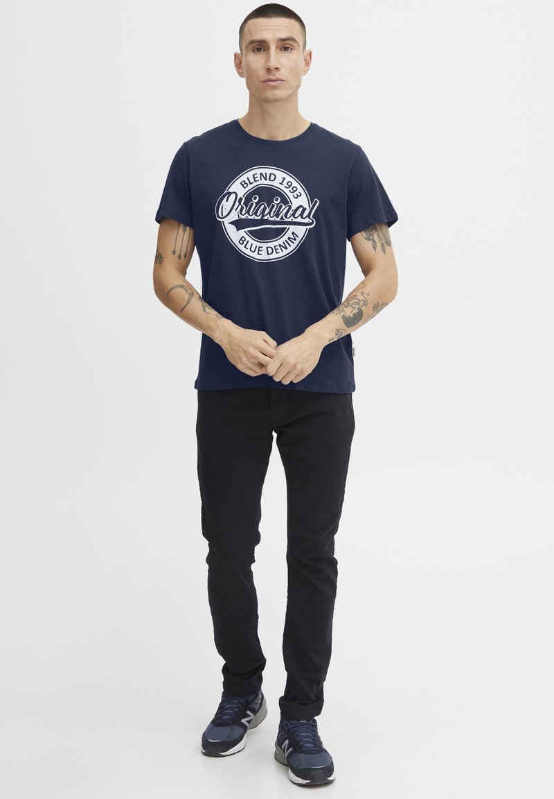 Mørkeblå bomull t-skjorte med hvit sirkulær "Original Blend 1993 Blue Denim" trykk, parret med svarte bukser og grå sneakers.