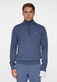 Miesten sininen neulepusero, jossa on korkea kaulus ja neljännesvetoketju. Varusteena ribattuja hihansuita ja helmaa sekä pieni valkoinen logo vasemmassa rinnassa.