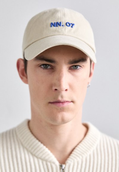 Beige bomullslue med blå "NN. 07" broderi foran, rund skjerm og avslappet passform. Brukt med en ribbestrikket genser.