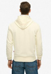 Sweat-shirt à capuche de couleur crème, à manches longues, en tissu texturé, avec poignets et ourlet côtelés, associé à un jean en denim bleu.