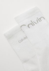 Socks de algodão brancos com um textura canelada e um logotipo "Calvin" cinza claro perto do topo. Design simples, sem padrões ou acentos adicionais.