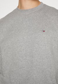 Szara bluza z okrągłym dekoltem o miękkiej teksturze, z małym logo w czerwonym, białym i niebieskim kolorze na lewej stronie klatki piersiowej.