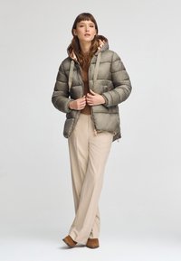 Puffjacka i olivgrön med huva, med dragkedjeslutning och mönstrad foder, matchad med beiga byxor och bruna ankelstövlar.