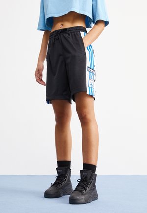 Person trägt schwarze Adidas Mesh-Shorts mit blauen Streifen und schwarze High-Top-Sneaker, steht auf hellblauem Boden vor weißer Wand.