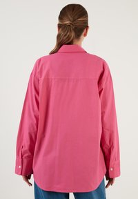 Pink, oversized button-up shirt with long sleeves and buttoned cuffs. Smooth texture, curved hem, and back yoke detailing.