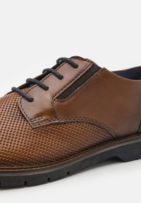 Brown leather shoe with perforated detailing, flat black sole, and dark laces. Features a smooth finish with contrasting textured panels.