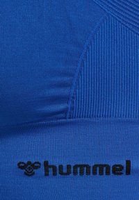 Blauwe sporttop met ribbelstructuur, soepele panelen en zwart Hummel-logo. Materiaal lijkt rekbaar en aansluitend voor optimale prestaties.