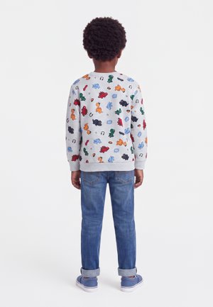 Enfant aux cheveux bouclés portant un sweat blanc avec des imprimés colorés de dinosaures, un jean bleu et des baskets bleues, debout de dos.