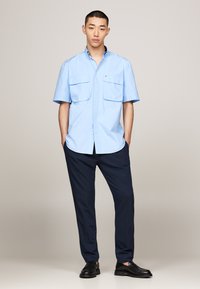 Light blue short-sleeve shirt with two front pockets, paired with navy trousers and black shoes, standing against a neutral background.