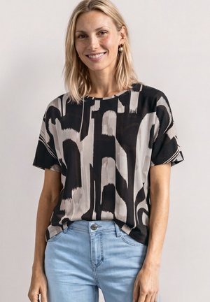 Kobieta o blond włosach sięgających ramion, ubrana w czarną i beżową koszulkę z abstrakcyjnym wzorem na krótkich rękawach oraz jasnoniebieskie dżinsy, uśmiechająca się.
