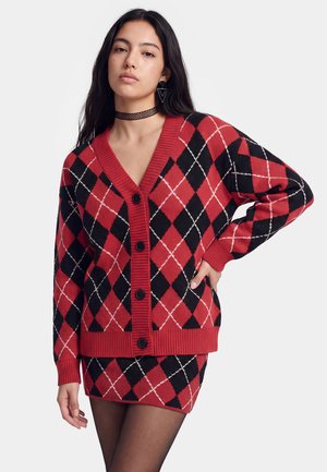 Vrouw met lang donker haar die een rood-zwart argyle vest en bijpassende rok draagt, zwarte panty, driehoekige oorbellen en een choker ketting.
