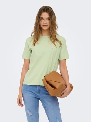 Ung kvinde iført lysegrøn t-shirt og slidte blå jeans, der holder en stor foldet brun læderclutch.