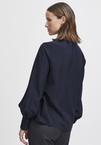 Marineblauwe blouse met lange, opbollende mouwen en geknoopte manchetten; gebroken stof met een geplooide halslijn; losvallend ontwerp.