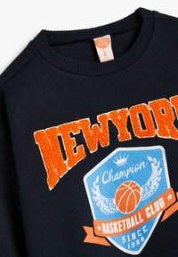 Sudadera de marina con el texto "NEW YORK" en tela de rizo naranja. Presenta un diseño de insignia en azul con un baloncesto y alas, etiquetado como "Champion Basketball Club."