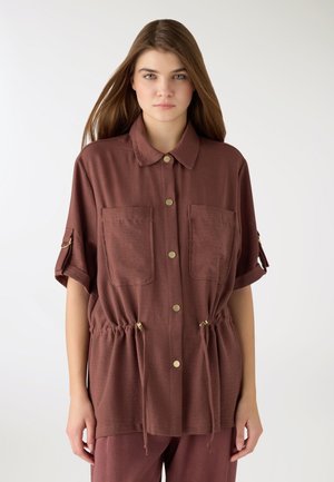 Giovane donna con capelli castani lisci indossa una camicia marrone larga con bottoni, due tasche sul petto e coulisse regolabili in vita, in piedi davanti a uno sfondo bianco.