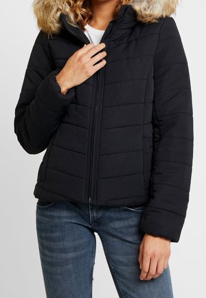 Personne portant une veste matelassée noire avec une capuche bordée de fourrure, fermée par une fermeture éclair, associée à un jean bleu sur un fond uni.