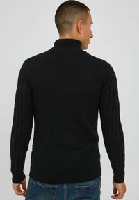 Pull noir en maille, avec un col haut, des poignets côtelés et un motif en tricot torsadé texturé sur les manches. Porté avec un jeans bleu.