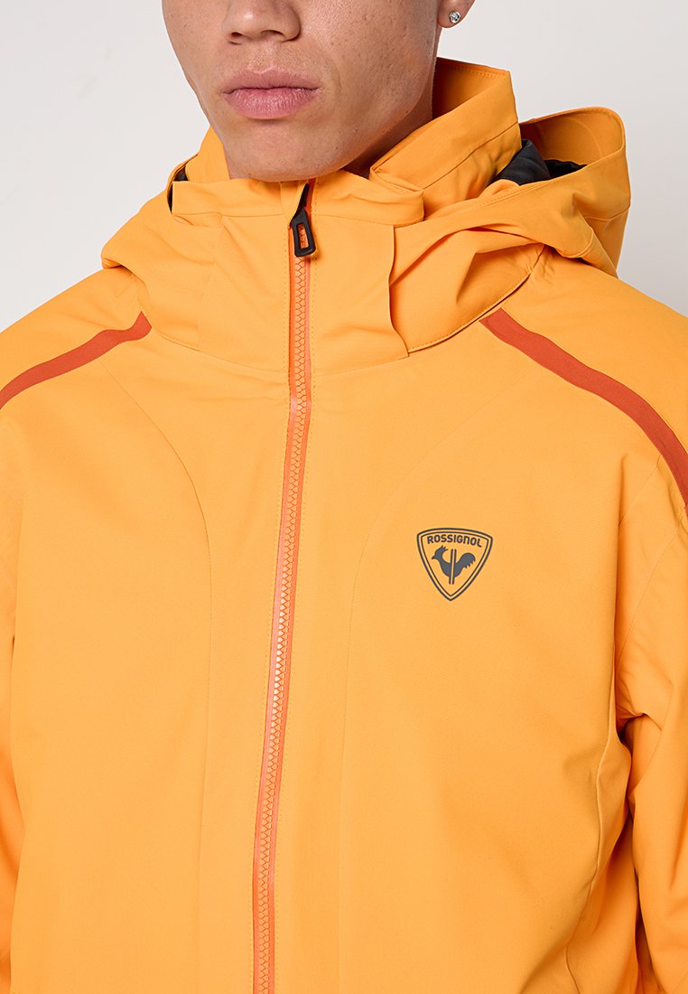 Żółta wodoodporna kurtka z wysokim kołnierzem, zamek błyskawiczny z przodu i kontrastowymi czerwonymi akcentami. Posiada logo na klatce piersiowej i gładką teksturę.