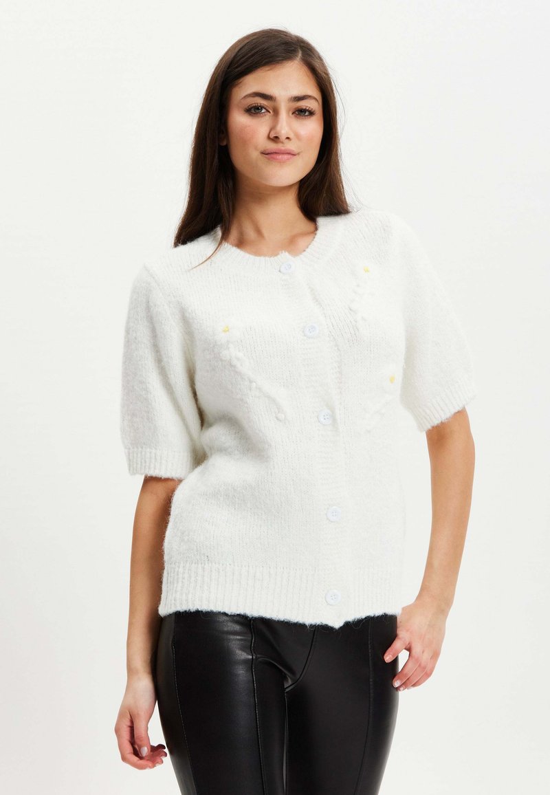 White, textured short-sleeve cardigan with a round neckline, featuring three buttons and subtle yellow detailing. Worn over black leather pants.