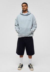  Lichtblauwe oversized hoodie met een gestructureerde afwerking en voorzak, gecombineerd met zwarte shorts tot de knie en zwarte sneakers met witte accenten.