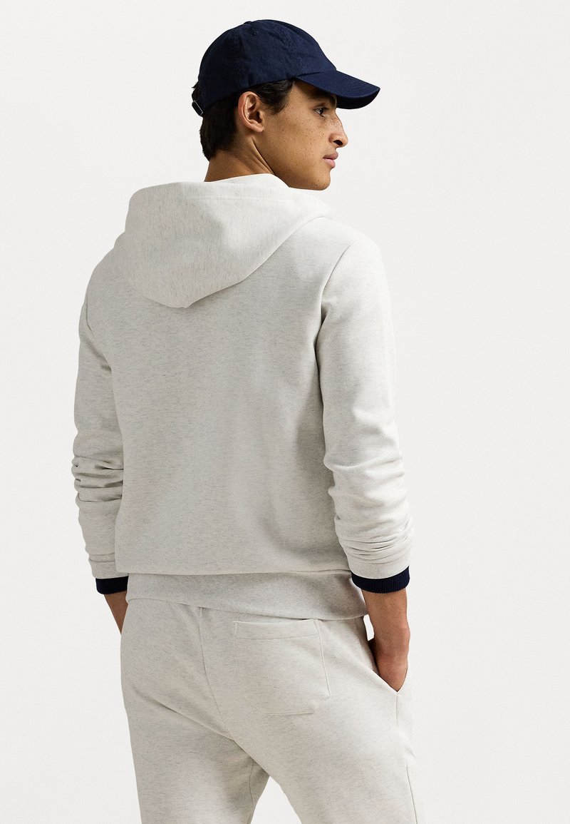 Sudadera gris claro con un bolsillo frontal, puños elásticos y capucha con cordón, llevada con pantalones a juego, complementada por una gorra azul marino.