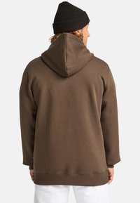 Brauner Kapuzenpullover mit entspannter Passform, gerippten Bündchen und Saum. Verfügt über eine große Kapuze und eine glatte Textur. Getragen mit einer schwarzen Mütze.