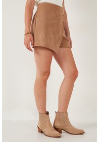 Shorts en daim de couleur beige avec un design enveloppant, comportant deux boutons et une texture lisse. Associés à des bottines beiges.