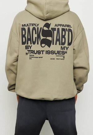 Beiger Hoodie mit großem schwarzem Text auf dem Rücken: "BACKSTAB'D by my 'TRUST ISSUES'" und kleinerem Text, der auf eine Streetwear-Marke verweist.