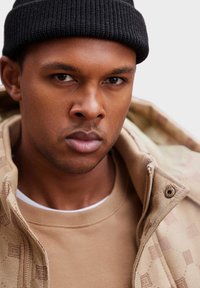 Béžová mikina s logem, vrstvená pod vzorovanou béžovou bundou; model má na sobě černou pletenou čepici, neutrální tón pleti, přímý pohled.