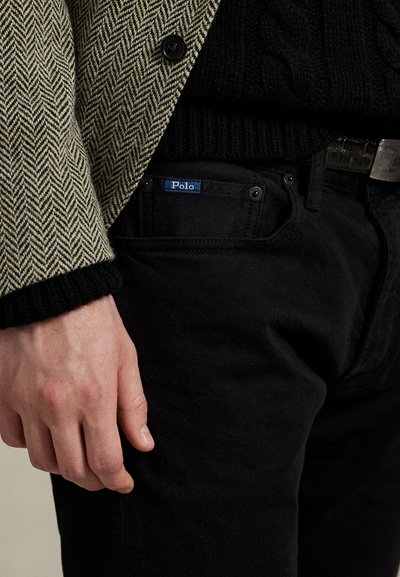 Svarte denimbukser med en synlig blå "Polo"-tag, kombinert med en teksturert svart strikkegenser og en jakke med sildebensmønster.