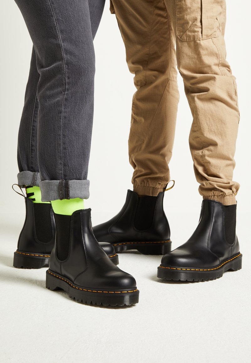 Deux personnes portent des bottines Chelsea en cuir noir avec des coutures jaunes, l'une en jean gris retroussé et chaussettes néon, l'autre en pantalon cargo beige.