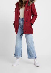 Röd parka med konstgjord päls på huva, med knäppningar och dragsko i midjan. Bärs över en krämfärgad topp och ljusblå vida jeans med vita sneakers.