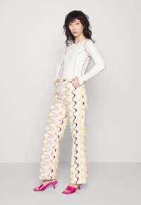 Top branco de manga longa com costura roxa, combinado com calças largas de padrão ondulado colorido. Modelo usa sandálias de salto rosas.