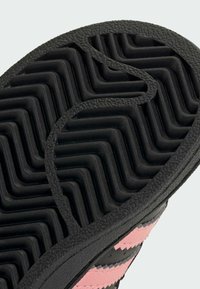 Semelle en caoutchouc noir avec un motif de crampons en zigzag et une surface texturée. Présente des accents supérieurs en rose et noir avec des coutures visibles.