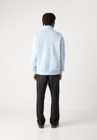 Chaqueta de pullover azul claro con un cuello alto, puños y dobladillo acanalados, combinada con pantalones rectos gris oscuro y zapatos marrones.