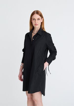 Frau mit glatten roten Haaren, die ein schwarzes Hemdkleid mit langen Ärmeln und Kragen trägt, steht mit einer Hand in der Tasche vor einem einfarbigen Hintergrund.