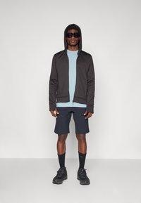 Felpa nera con zip e cappuccio, t-shirt azzurra chiara, pantaloni corti scuri, calzini neri e scarpe robuste nere. Tessuti lisci e aderenti.