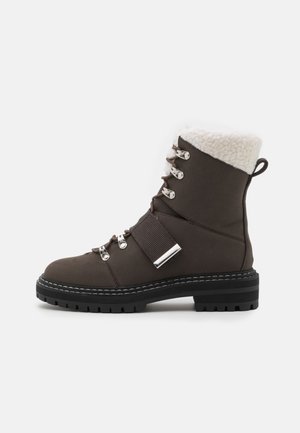 Botte d'hiver marron avec semelle noire épaisse, œillets métalliques, sangle à fermeture auto-agrippante et doublure en polaire blanche au niveau du col de la cheville, vue latérale.