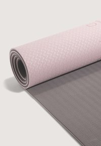 Joga prostirka u ružičastoj boji s teksturiranom površinom; dok je uvučena, otkriva kontrastnu sivu donju stranu s valovitom šablonom.