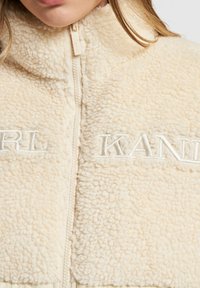 Beige fleecejacka med hög krage, hel dragkedja och broderade "KARL" och "KANE" framtill. Mjuk textur och varm design.