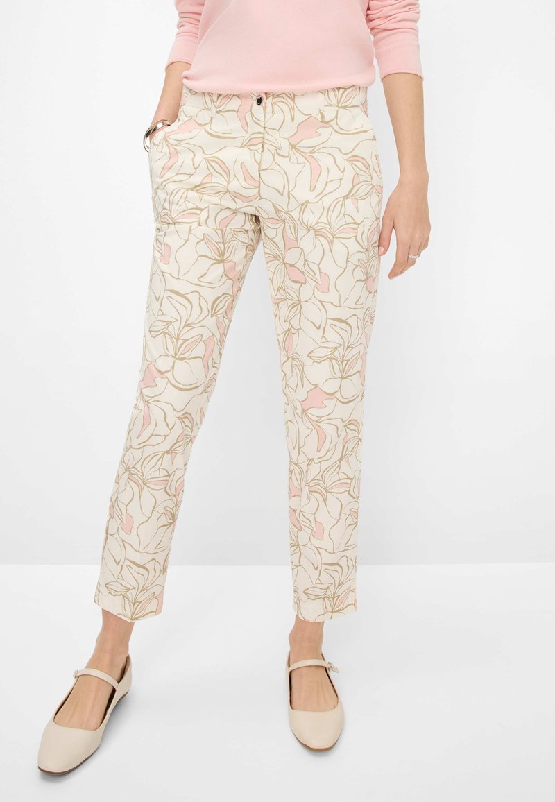 Personne portant un pantalon beige à imprimé floral rose et vert, un haut à manches longues rose clair, des ballerines Mary Jane beiges, les mains reposant le long du corps.
