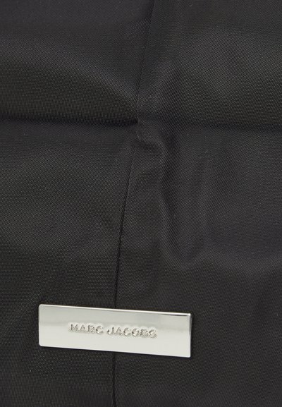 Sac en nylon noir avec une étiquette rectangulaire en métal argenté embossée avec "MARC JACOBS". La texture est lisse avec un motif de tissage subtil.
