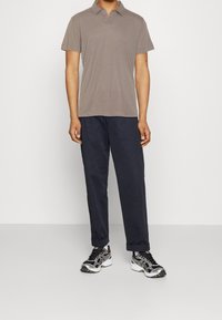 Brun poloshirt med korte ærmer, kombineret med navy bukser og sorte sneakers med sølvdetaljer, stående mod en ensfarvet baggrund.