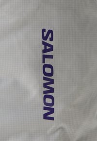 Szürke szövet texturált felülettel, amelyen nagy, vastag lila betűkkel szerepel a "SALOMON" felirat. Minimalista dizájn, amely a márkázást helyezi előtérbe.