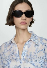Occhiali da sole neri con montatura ovale abbinati a una blusa bianca trasparente con un motivo floreale blu e un colletto classico.