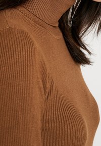 Pull en maille côtelée marron avec une silhouette ajustée. Texture douce et col haut. Le motif présente des côtes verticales sur toute la surface.
