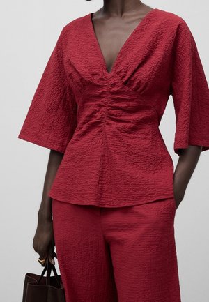 Femme portant un haut rouge texturé à col en V avec fronces au centre et un pantalon assorti, tenant un sac à main marron foncé dans une main.